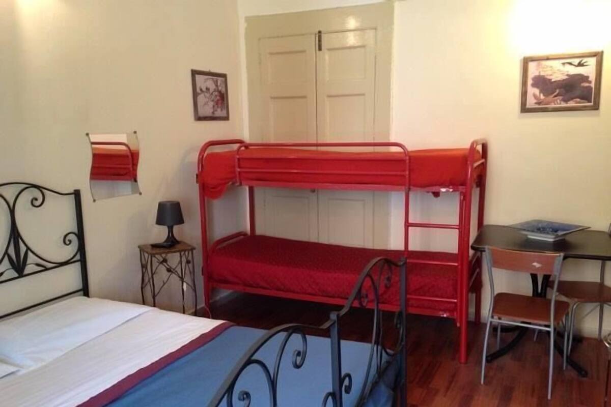 A dormitory room with bunk beds, a desk, a chair, a small table with a lamp, and a door.