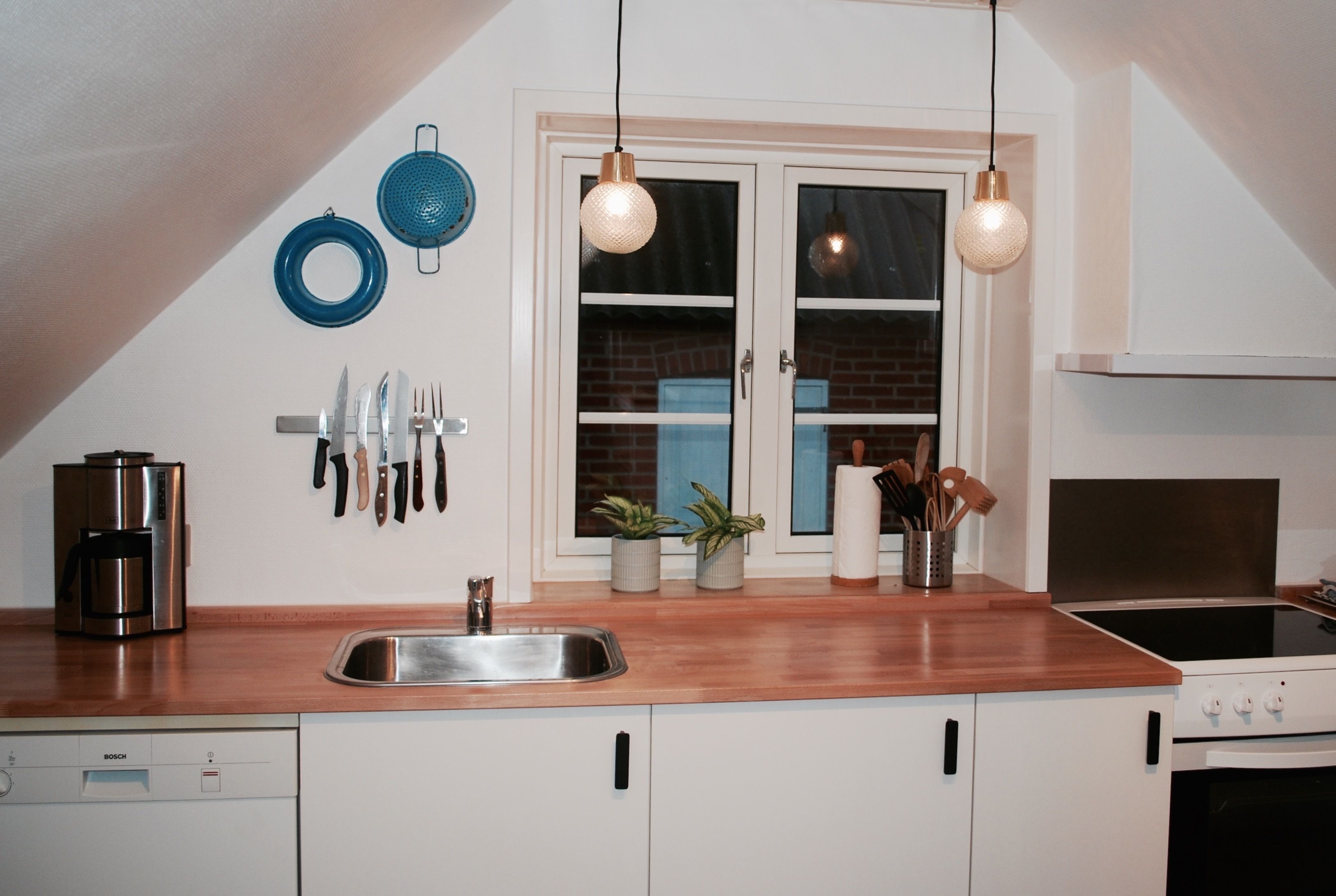 A kitchen with a sink, stove, and various utensils. There are two hanging lights and a window with a view of a brick building.