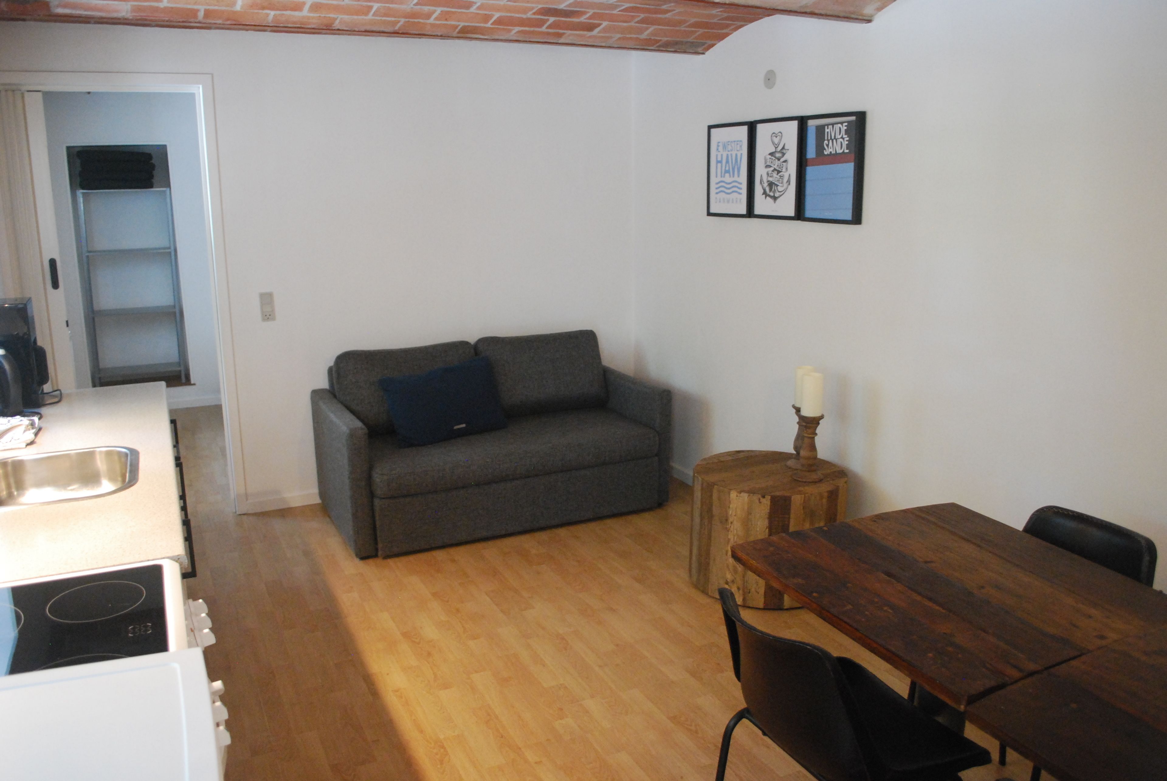Ein kompakter Wohnbereich mit grauem Sofa, Holzesstisch und einem kleinen, runden Hocker. In der Ecke ist ein weißes Gerät zu sehen, und an der Wand hängen gerahmte Bilder.