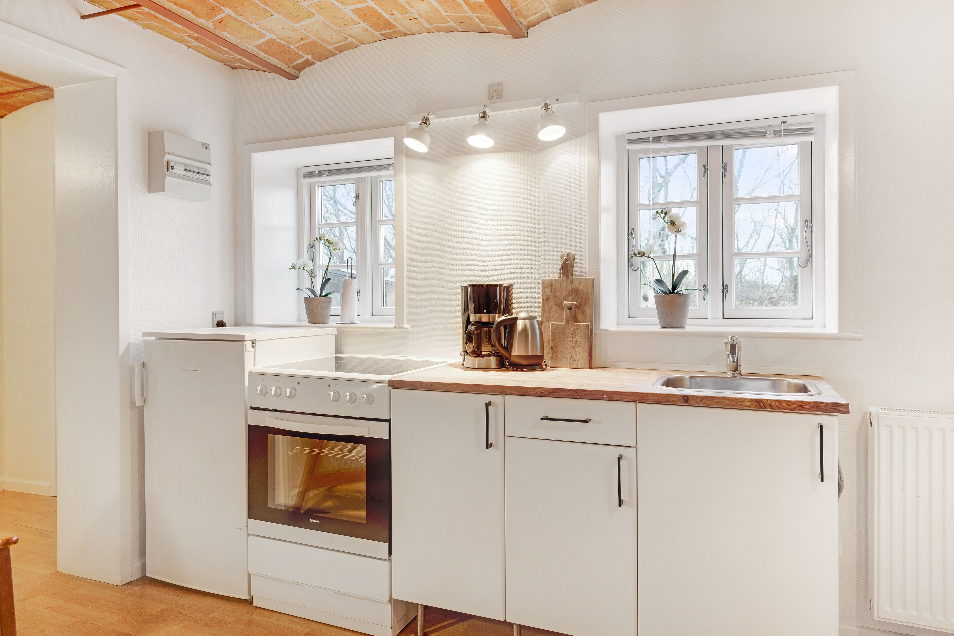 A compact kitchen with white cabinetry, a built-in oven, a coffee maker, and two windows with potted plants.