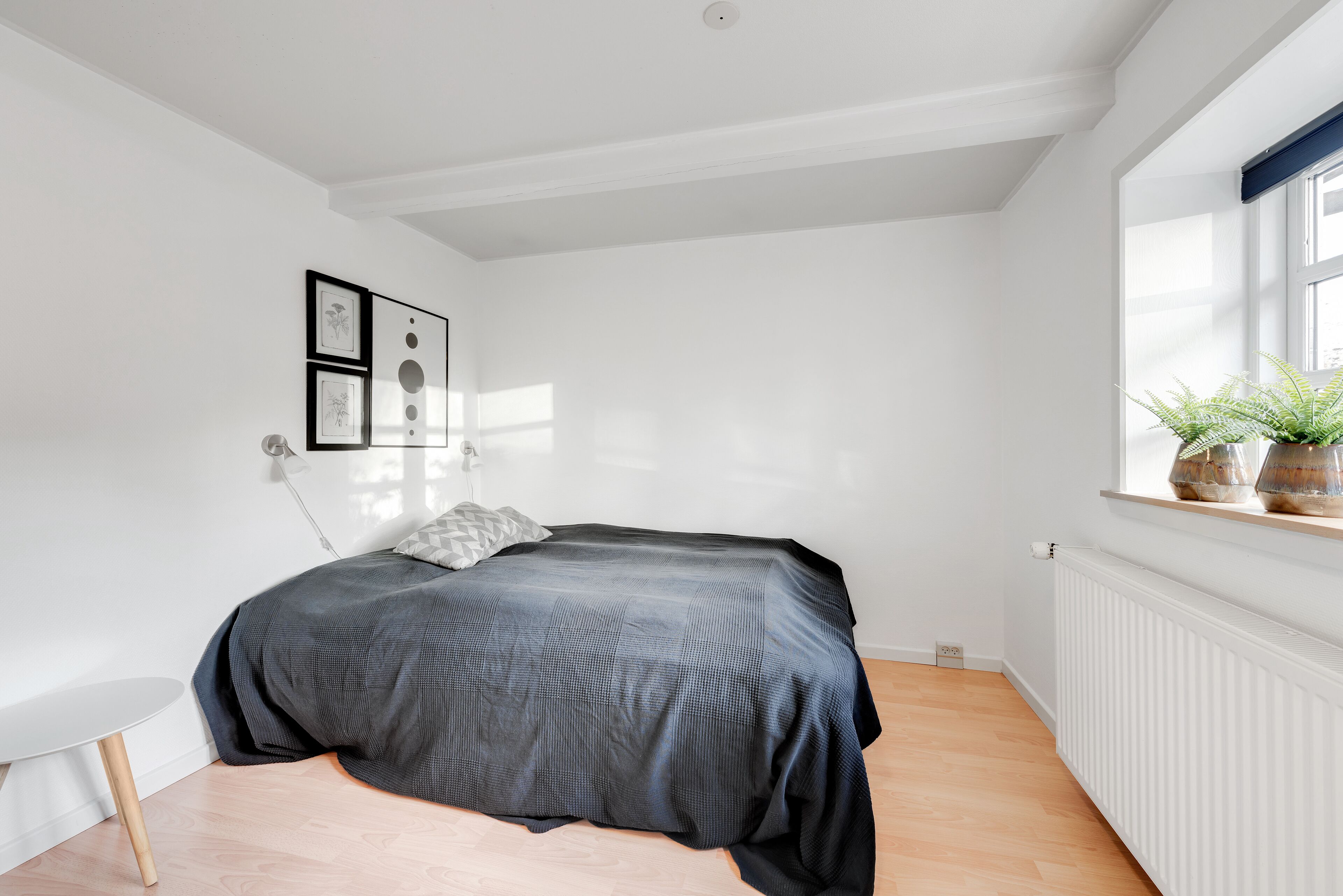 Un dormitorio con una cama, una mesita, un radiador y una ventana con una planta.