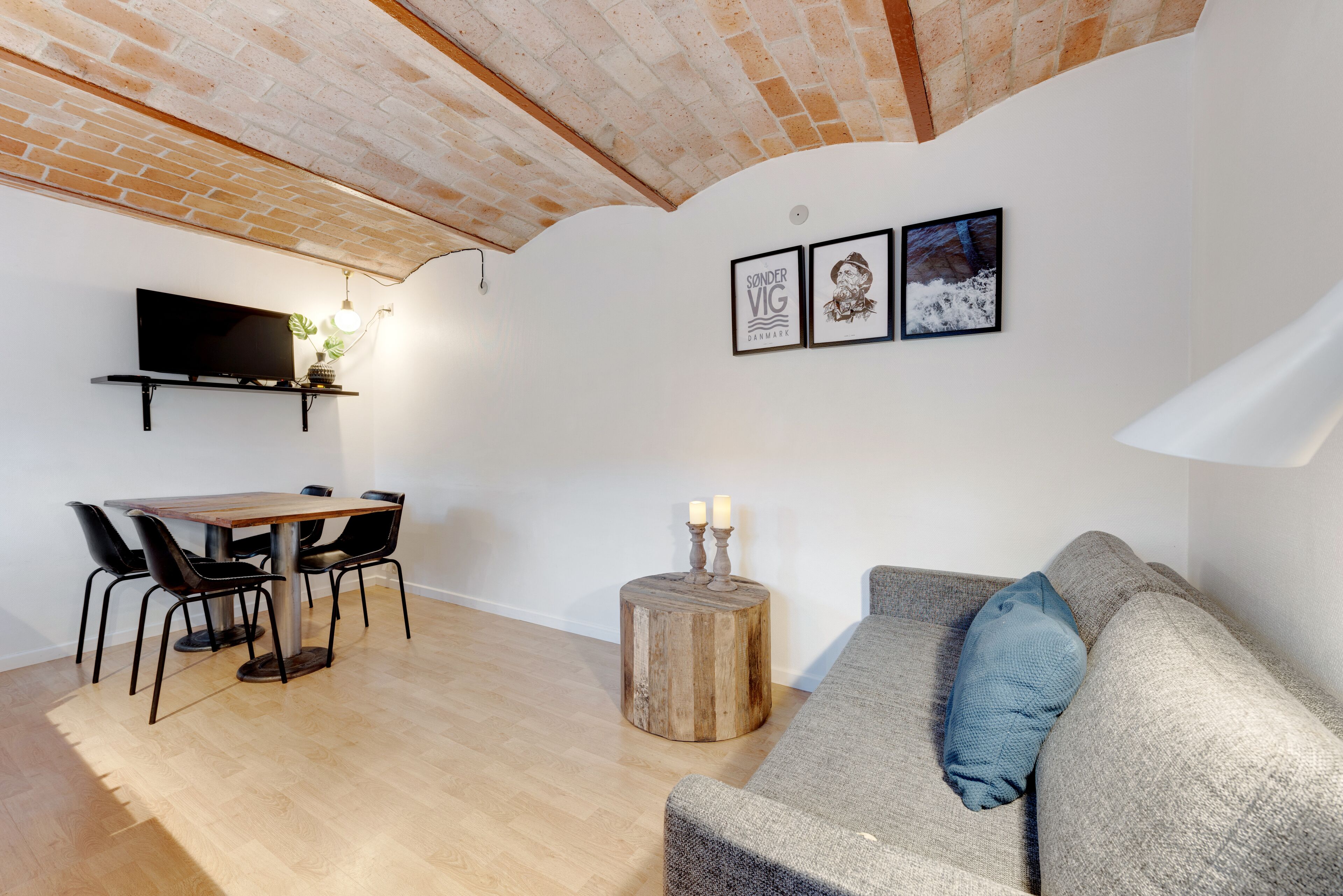 A cozy living area with a wooden dining table, black chairs, a grey sofa with a blue pillow, and a round wooden side table with candles.