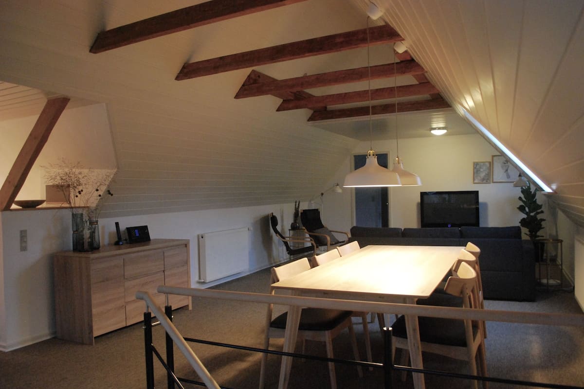 Ein modernes Loft mit freiliegenden Holzbalken, einem Essbereich mit Tisch und Stühlen, einem Wohnzimmer mit Sofa und einer Küchenzeile inklusive Kühlschrank und Schränken.