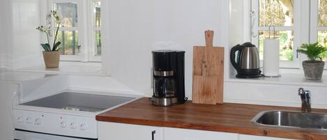 A kitchen with white cabinets, a wooden countertop, a coffee maker, a kettle, and a window with a view of trees.