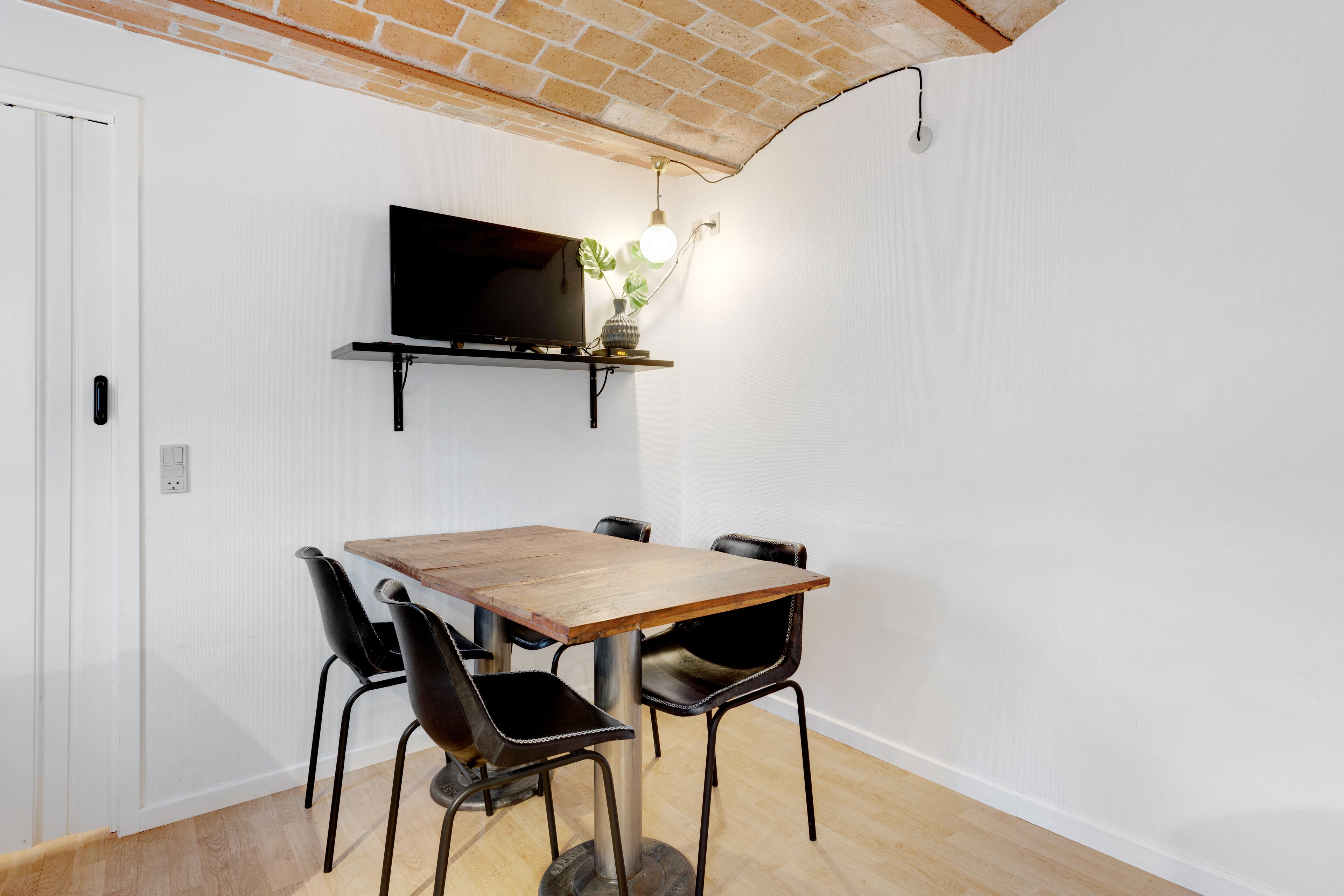A dining area with a wooden table and four chairs, a flat-screen TV mounted on the wall, and a hanging light fixture.
