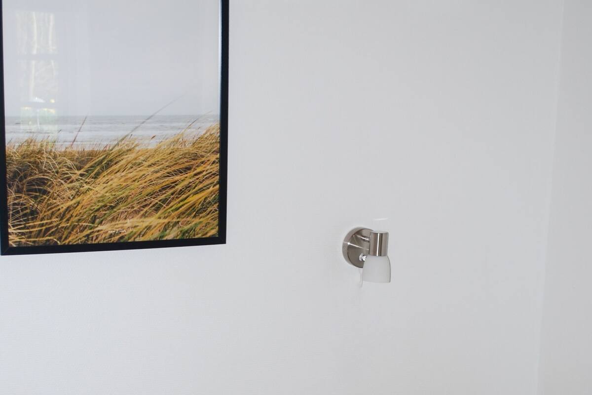 Ein Zimmer mit zwei Betten, einem kleinen Hocker, einer Wandleuchte und einem Bilderrahmen mit einem Strandmotiv.