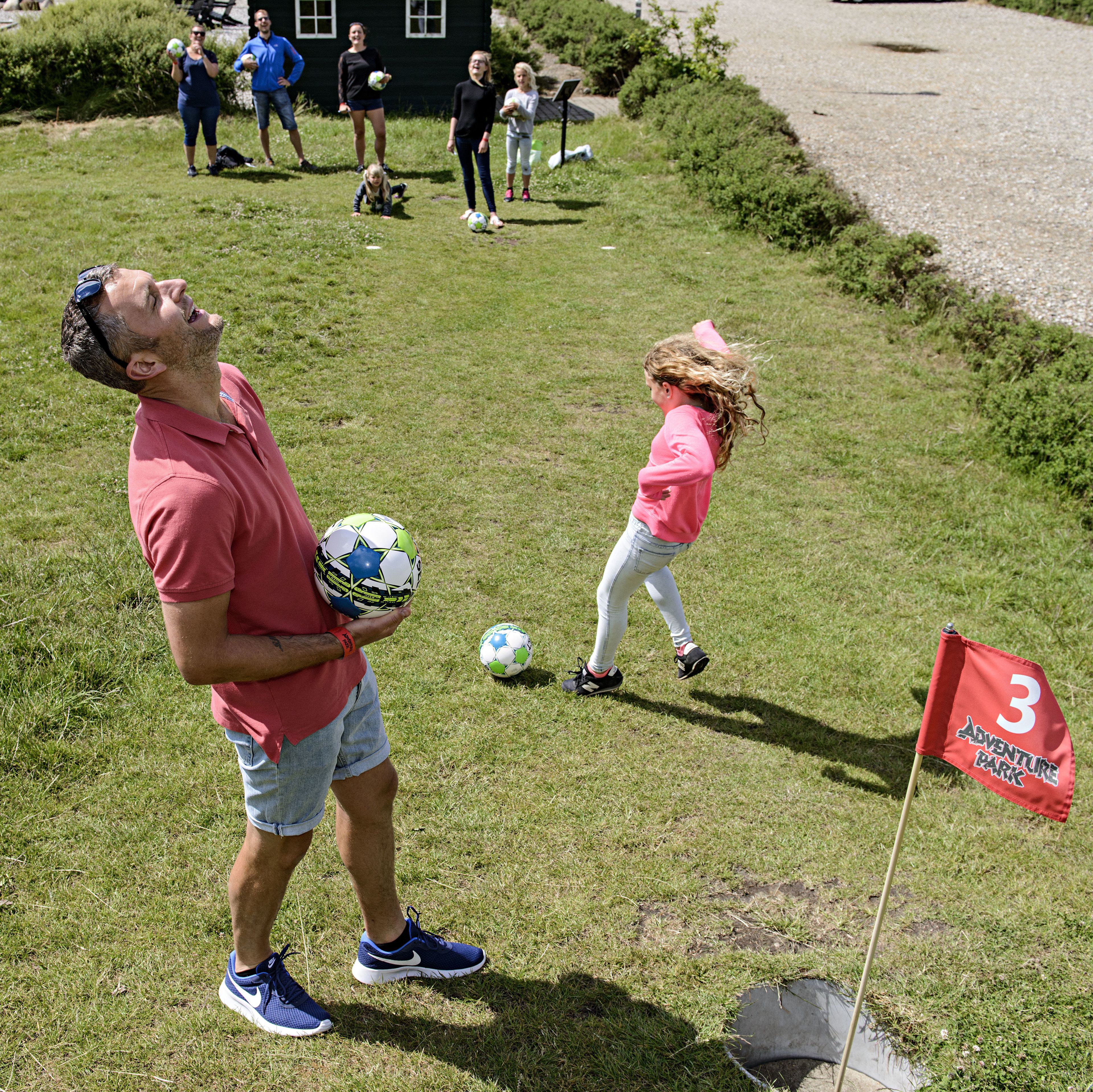 Un hombre con camisa roja y pantalones cortos azules sostiene un balón de fútbol, una niña con camisa rosa y jeans, y una bandera roja con el número 3 y la frase 