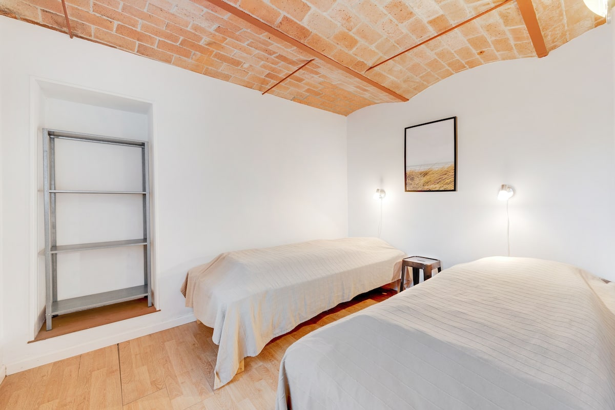 Ein Schlafzimmer mit zwei Betten, Holzboden, Ziegeldecke und einer weißen Wand mit einem Bild und Wandleuchten.