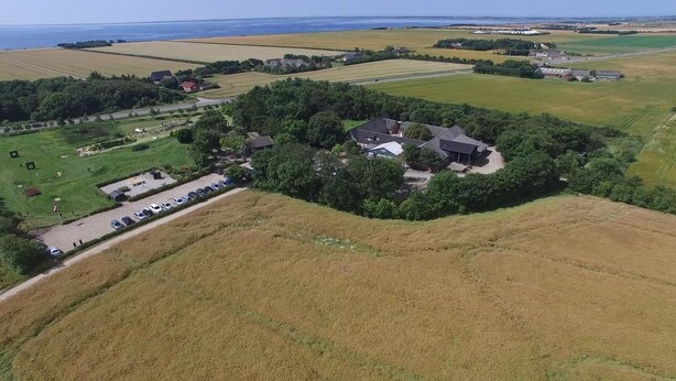 An aerial view of a rural area with a cluster of houses surrounded by fields, a parking lot, and a body of water in the distance. - Adventure Park Apartments (Ringkobing)