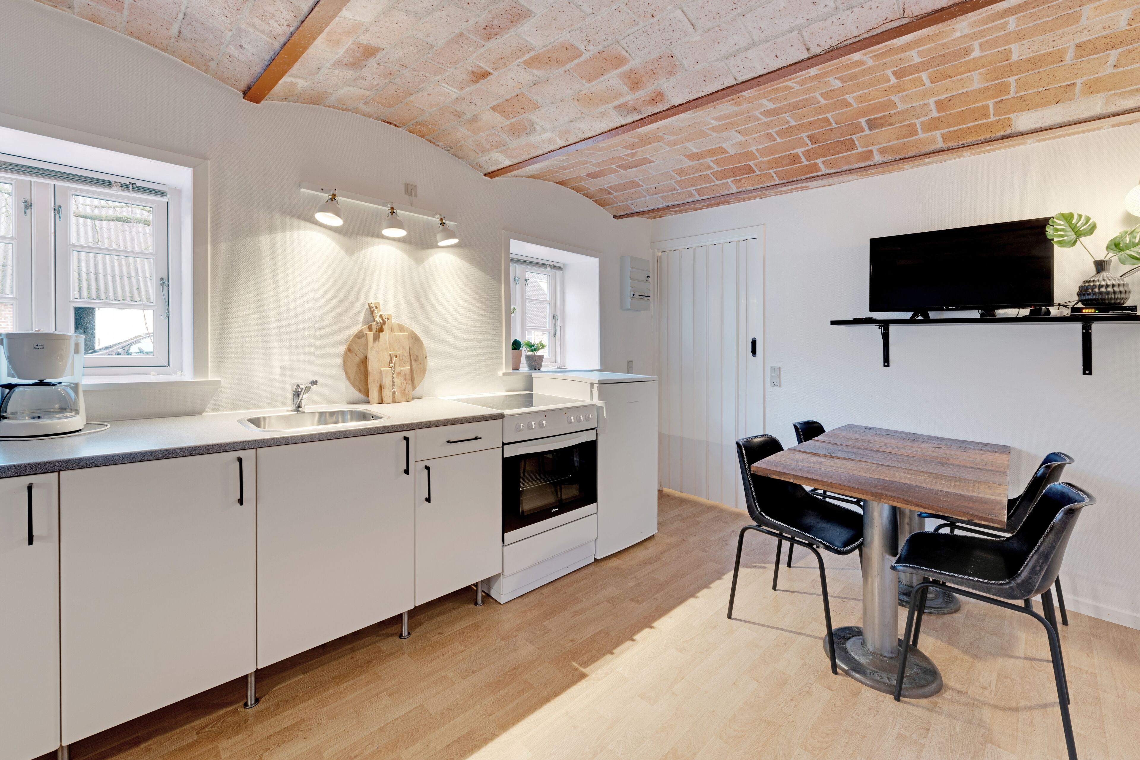A modern kitchen with white cabinets, a wooden dining table, and a TV mounted on the wall.