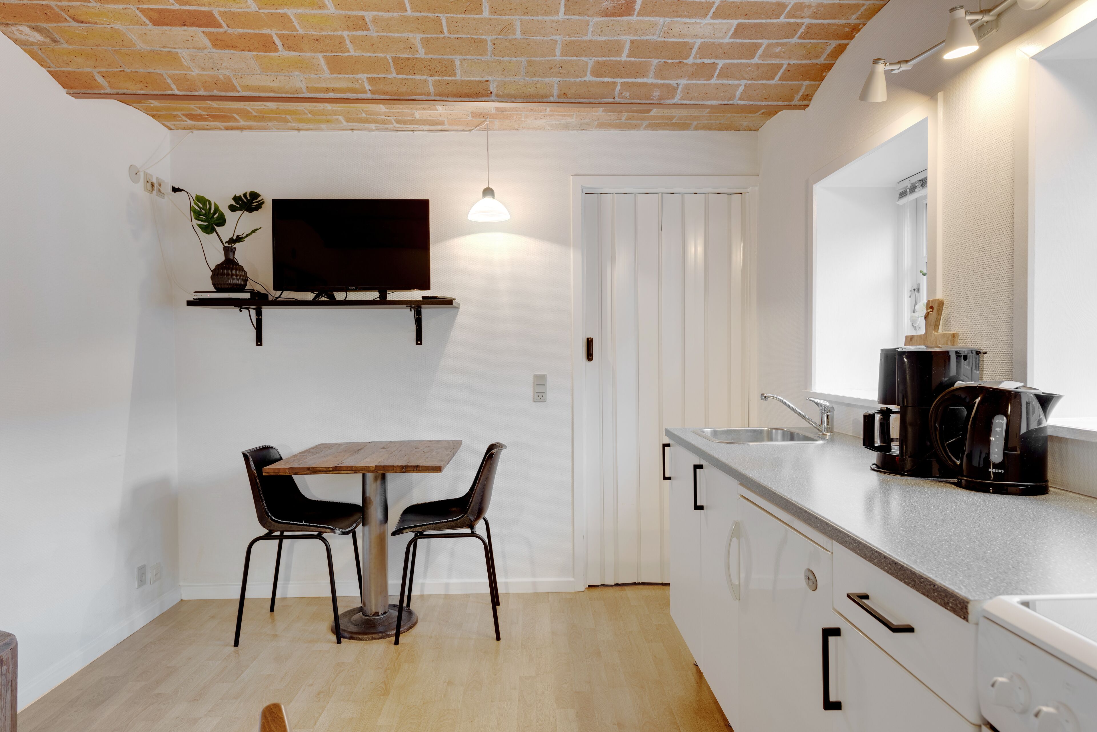 Eine kompakte Küche mit einem Holztisch, zwei Stühlen, einem eingebauten Ofen, einer Kaffeemaschine und einem an der Wand montierten Fernseher.