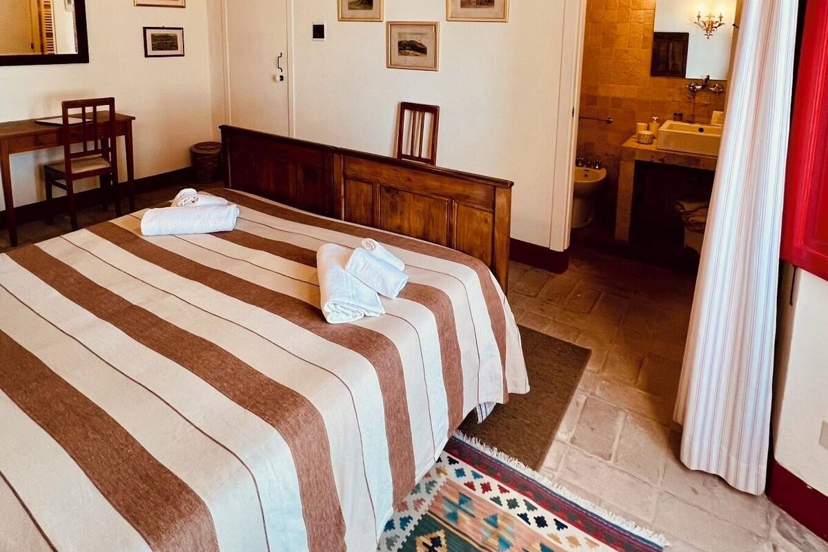 Ein Hotelzimmer mit einem großen Bett, einem Holztisch, einem Stuhl, einem kleinen Tisch, einem durch eine offene Tür einsehbaren Badezimmer und einem gemusterten Teppich.