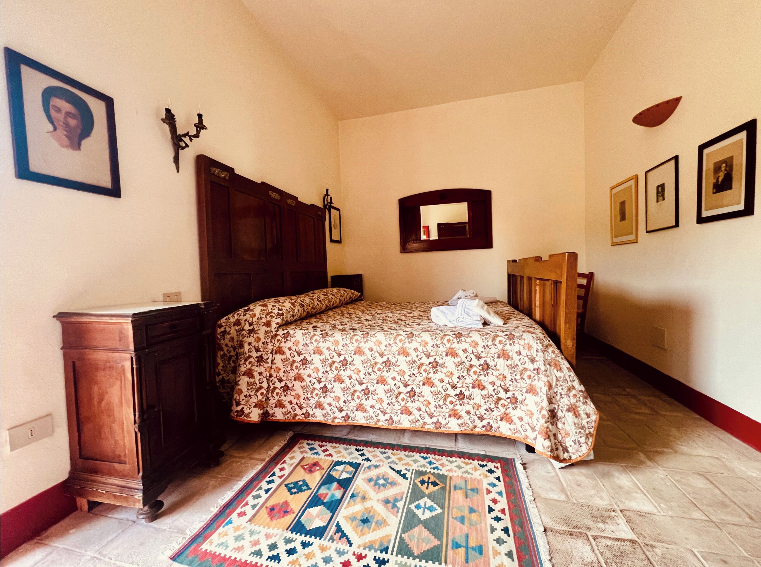 A bedroom with a wooden headboard, a bed with a floral bedspread, a patterned rug, a wooden nightstand, and a mirror on the wall.