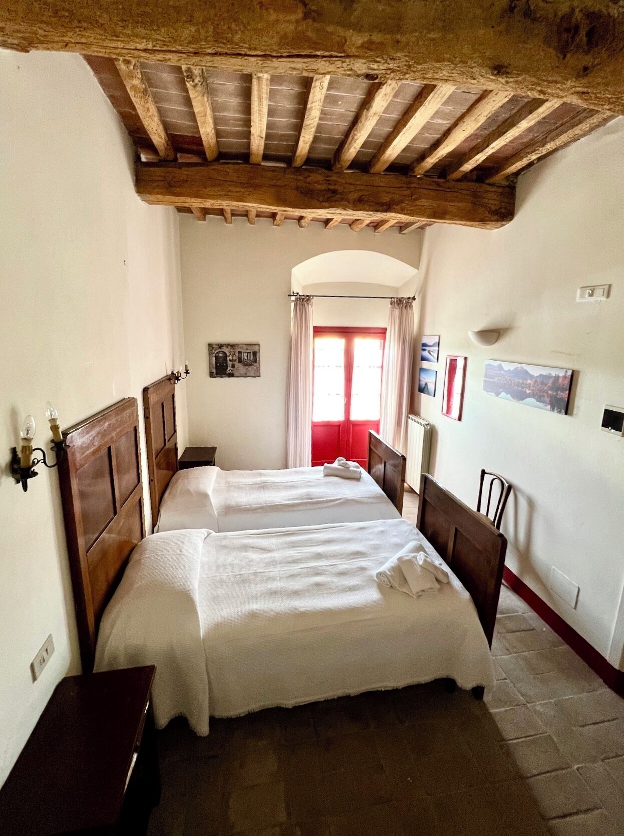 Ein Zimmer mit zwei Betten, Holzbalken an der Decke, einem Fenster mit roten Vorhängen und Wandbildern.