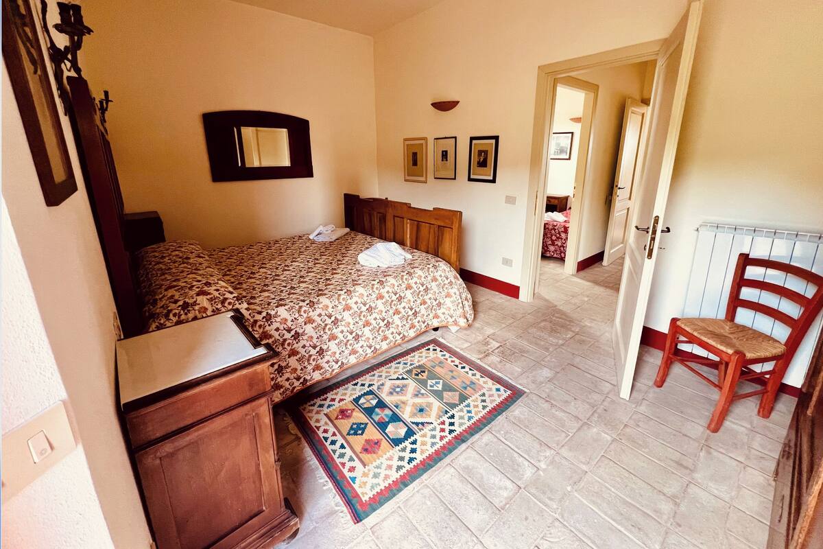Ein Schlafzimmer mit einem Bett, einer hölzernen Kommode, einem Stuhl, einem gemusterten Teppich und einem Flur, der in einen anderen Raum führt.