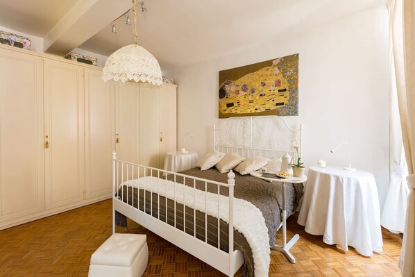 A bedroom with a white bed, a bedside table, a nightstand, a hanging lamp, and a painting on the wall.