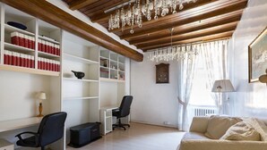 A room with wooden beams, a chandelier, a desk with a chair, a bookshelf, a couch, and a window with curtains. - Via Laurina 40 Apartment (Rome)