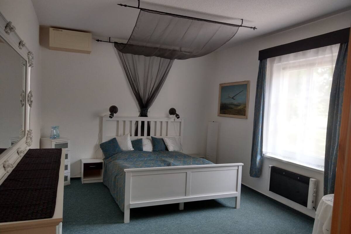 Un dormitorio con una cama blanca, una mesita de noche, un aparador, una ventana con cortinas y un radiador.