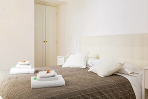 Un letto rifatto con cura, con una coperta a righe marroni, cuscini bianchi e asciugamani piegati con fiori arancioni su un comodino bianco.
