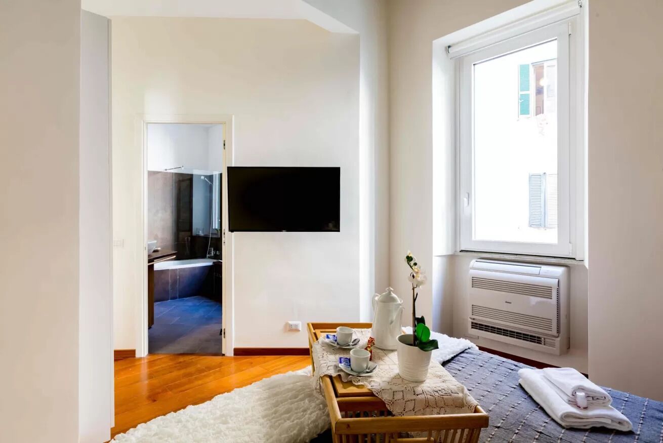 Une pièce moderne avec un plancher en bois, un canapé blanc, une petite table recouverte d’une nappe blanche, une plante en pot, un téléviseur à écran plat fixé au mur et une fenêtre avec un appareil de climatisation.