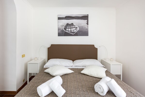 A neatly made bed with white pillows and rolled towels, a white bedside table with a lamp, and a framed picture of a boat on water above the bed. - A World Apart - Trevi Apartments (Rome)