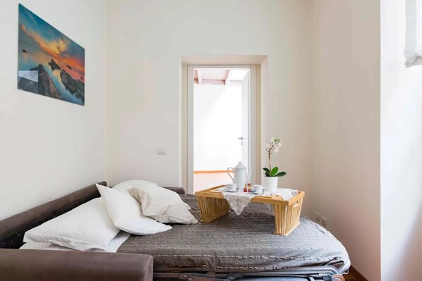 A small, cozy room with a bed, a bedside table with a teapot, and a window with a view of the outside.