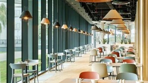 Um restaurante moderno com mesas e cadeiras compridas, grandes janelas e luminárias pendentes.