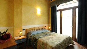 A bedroom with a wooden headboard, a floral bedspread, a nightstand with a lamp, and a view of the outdoors through a door. - Agriturismo Gian Galeazzo Visconti (Valeggio sul Mincio)