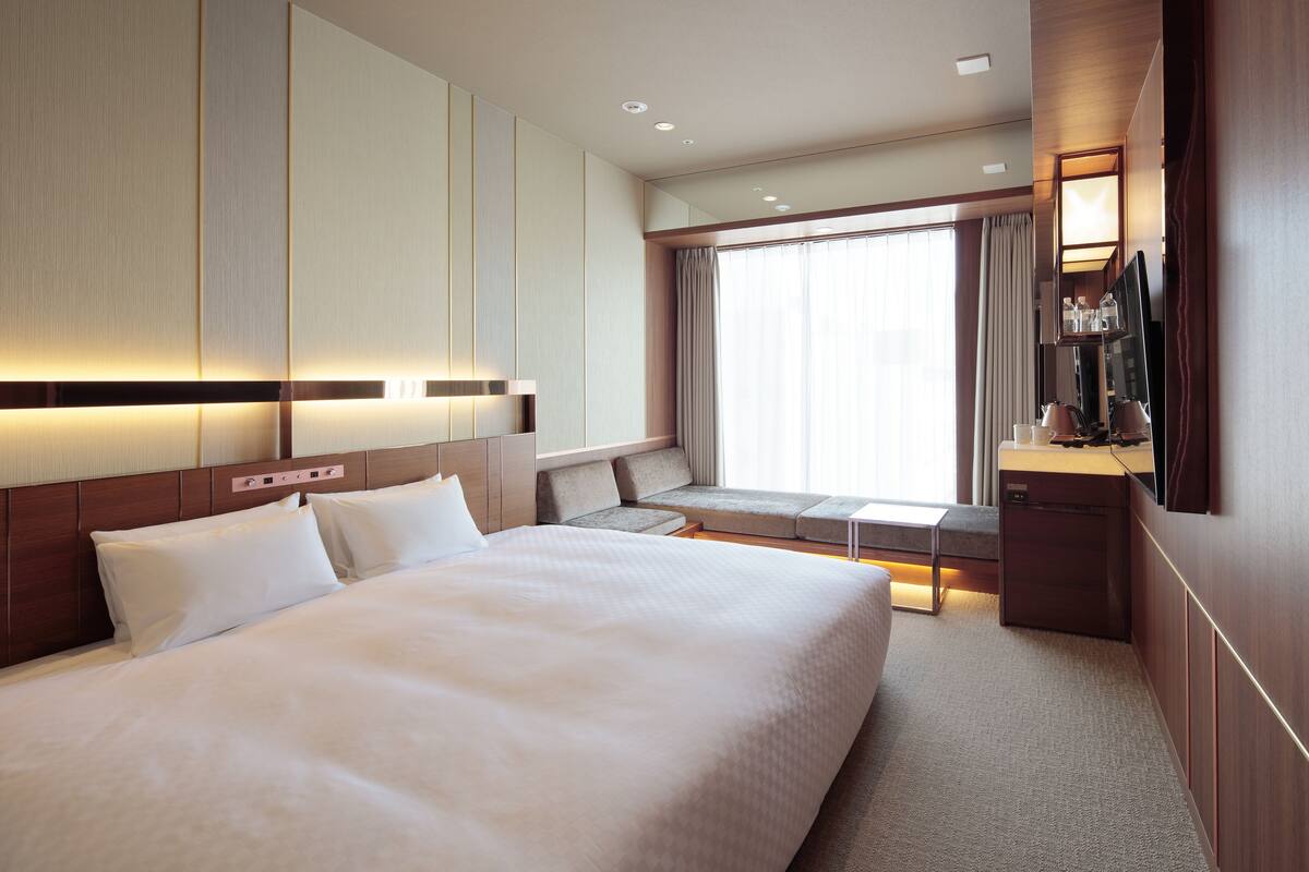 A hotel room with a large bed, a desk with a flat-screen TV, a seating area with a sofa and armchairs, and a large window with curtains.