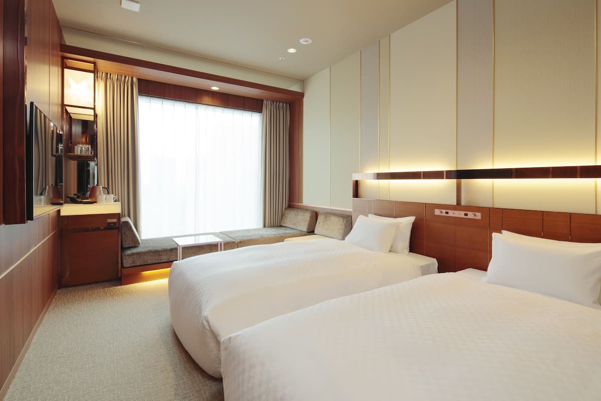 A hotel room with two beds, a desk with a lamp, a TV, and a large window with curtains.