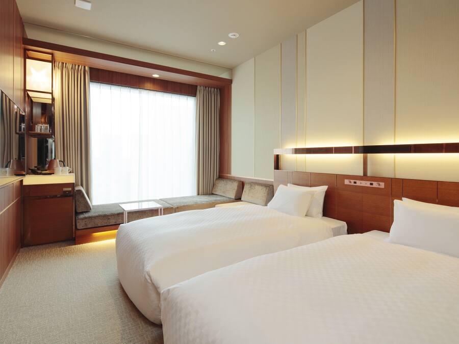 A hotel room with two beds, a desk with a lamp, a TV, and a large window with curtains.