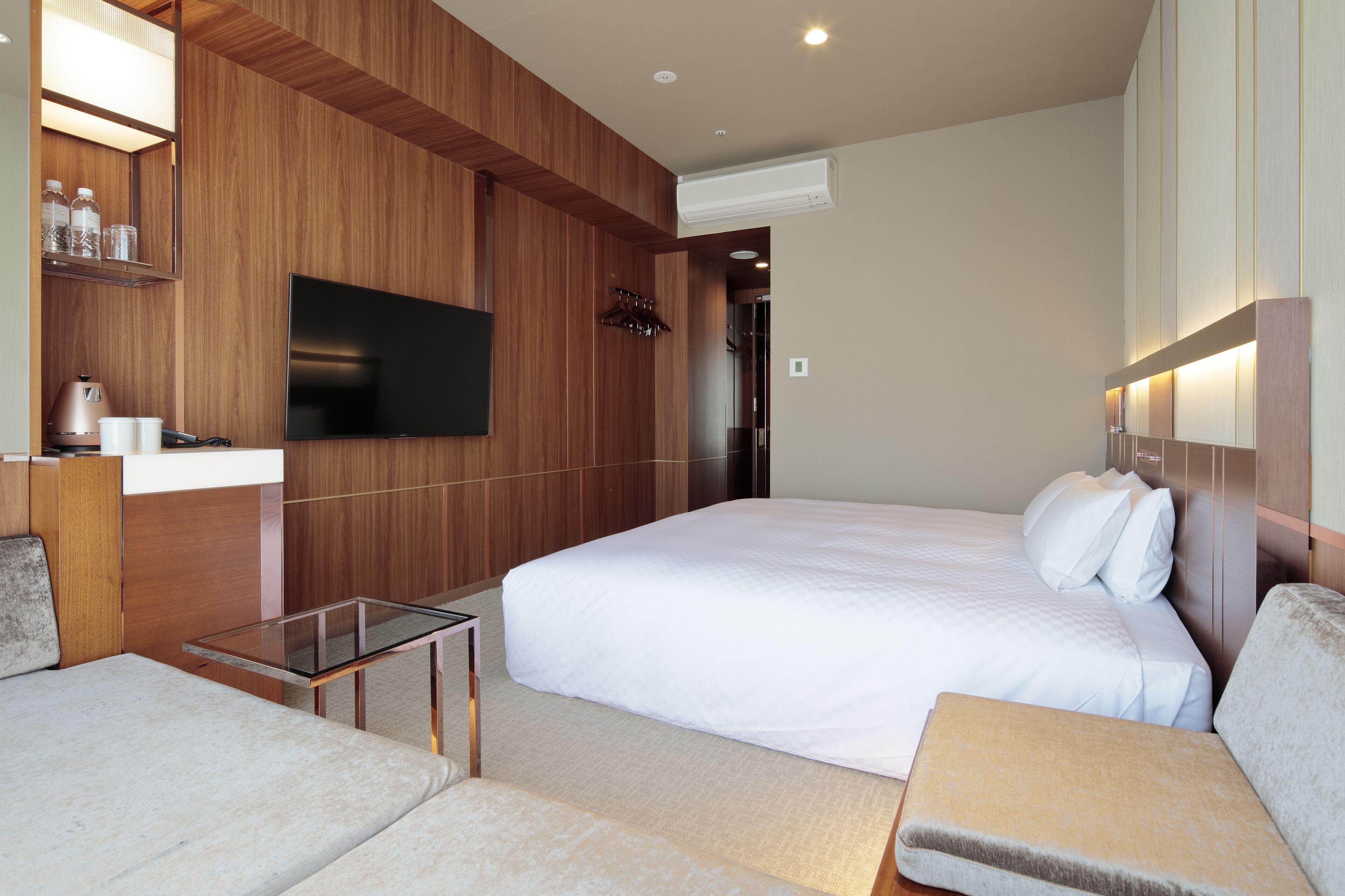 A modern hotel room with a large bed, a flat-screen TV mounted on the wall, a small table, and a sofa.