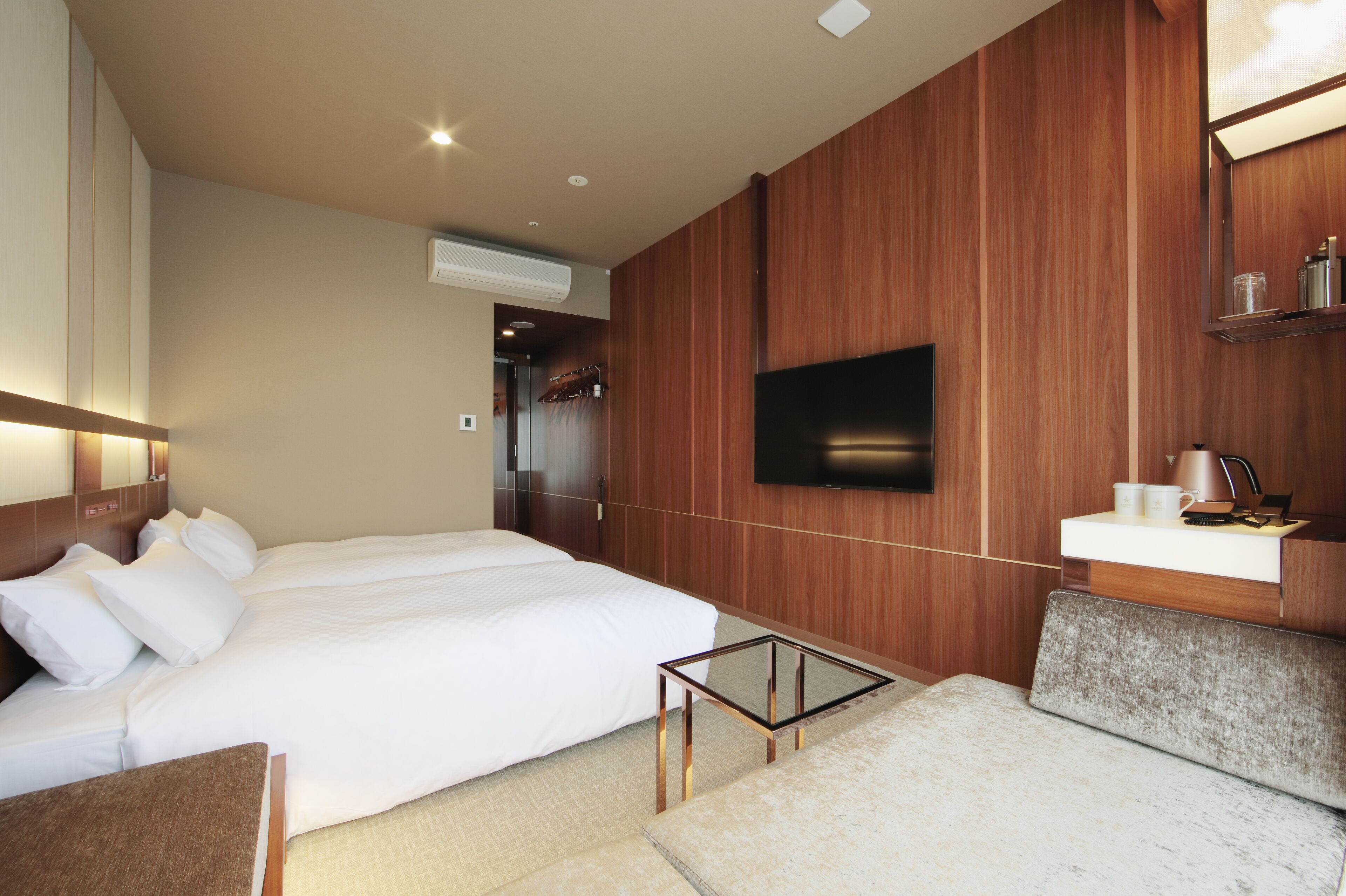Ein modernes Hotelzimmer mit einem großen Bett, einem flachbildfernseher an der Wand, einem kleinen Nachttisch und einer integrierten Schminkkommode mit Spiegel.