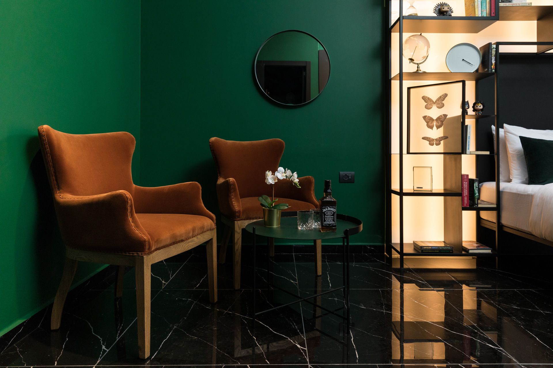 Una habitación con dos sillones de cuero marrón, una mesita redonda con una orquídea en maceta, una botella de whisky Jack Daniel's y una estantería con mariposas enmarcadas y libros.
