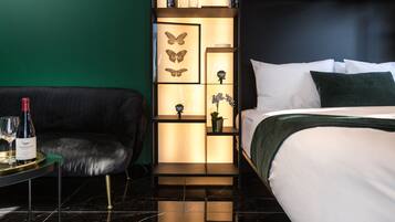 Ein modernes Hotelzimmer mit einer schwarzen Ledersofa, einem Bett mit weißer Bettwäsche, einem Nachttisch mit einer Weinflasche und einer dekorativen Wandverkleidung mit Schmetterlingskunst und Orchideen.