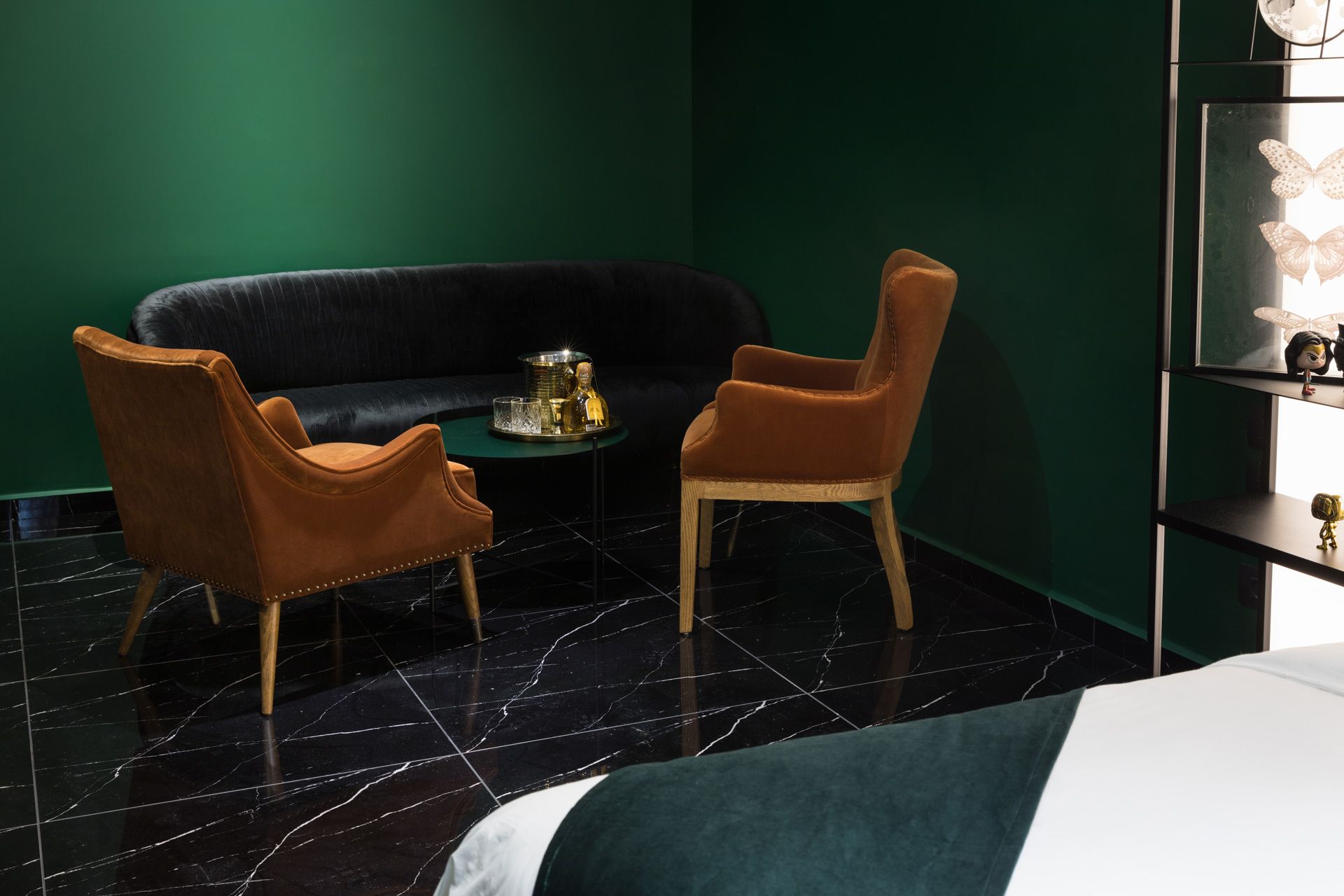 A room with a black sofa, two brown armchairs, a small round table with a gold tray, and a bed with a green and white duvet.