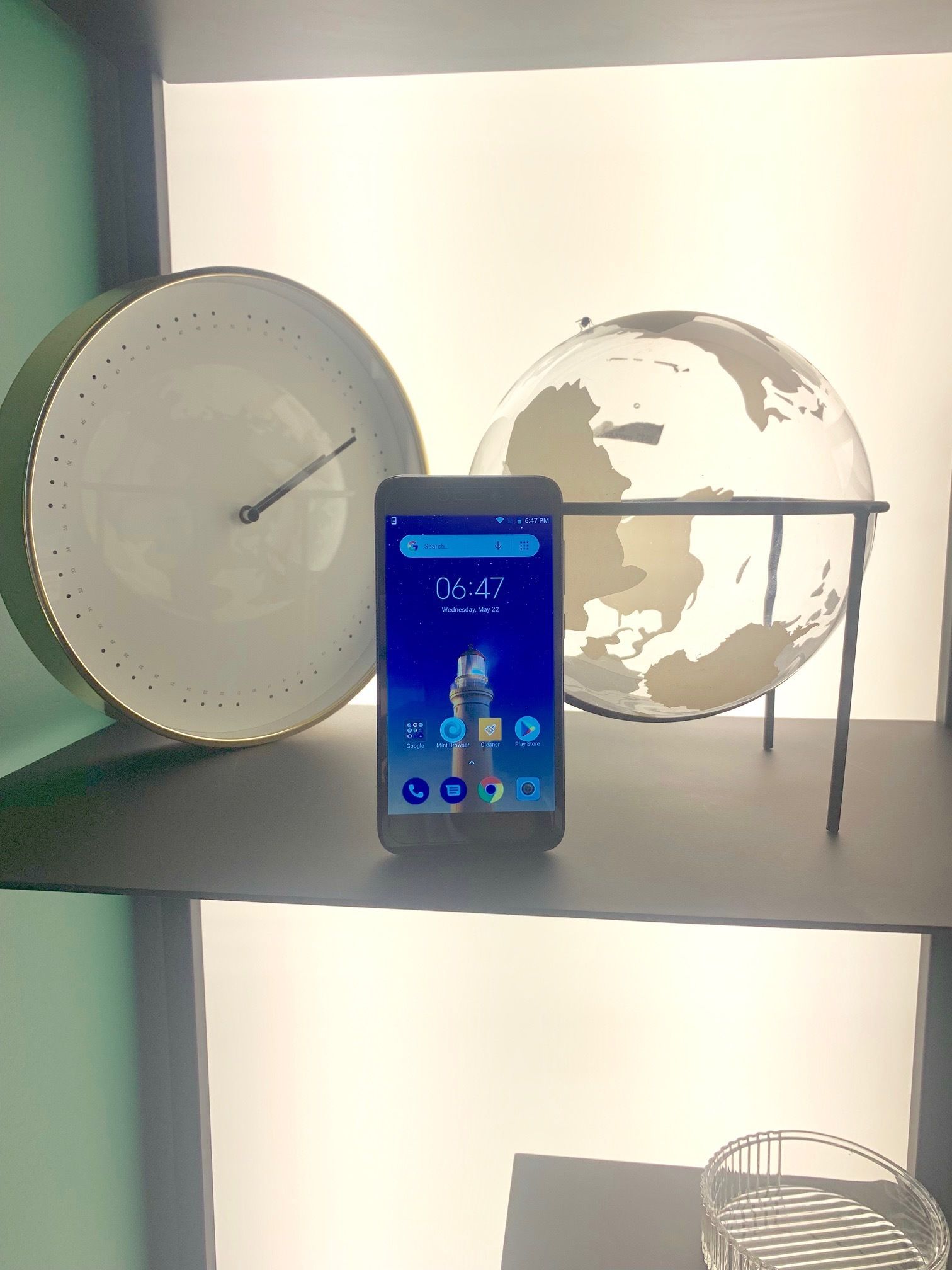 A smartphone displaying the time and date, a clock, and a globe on a shelf.