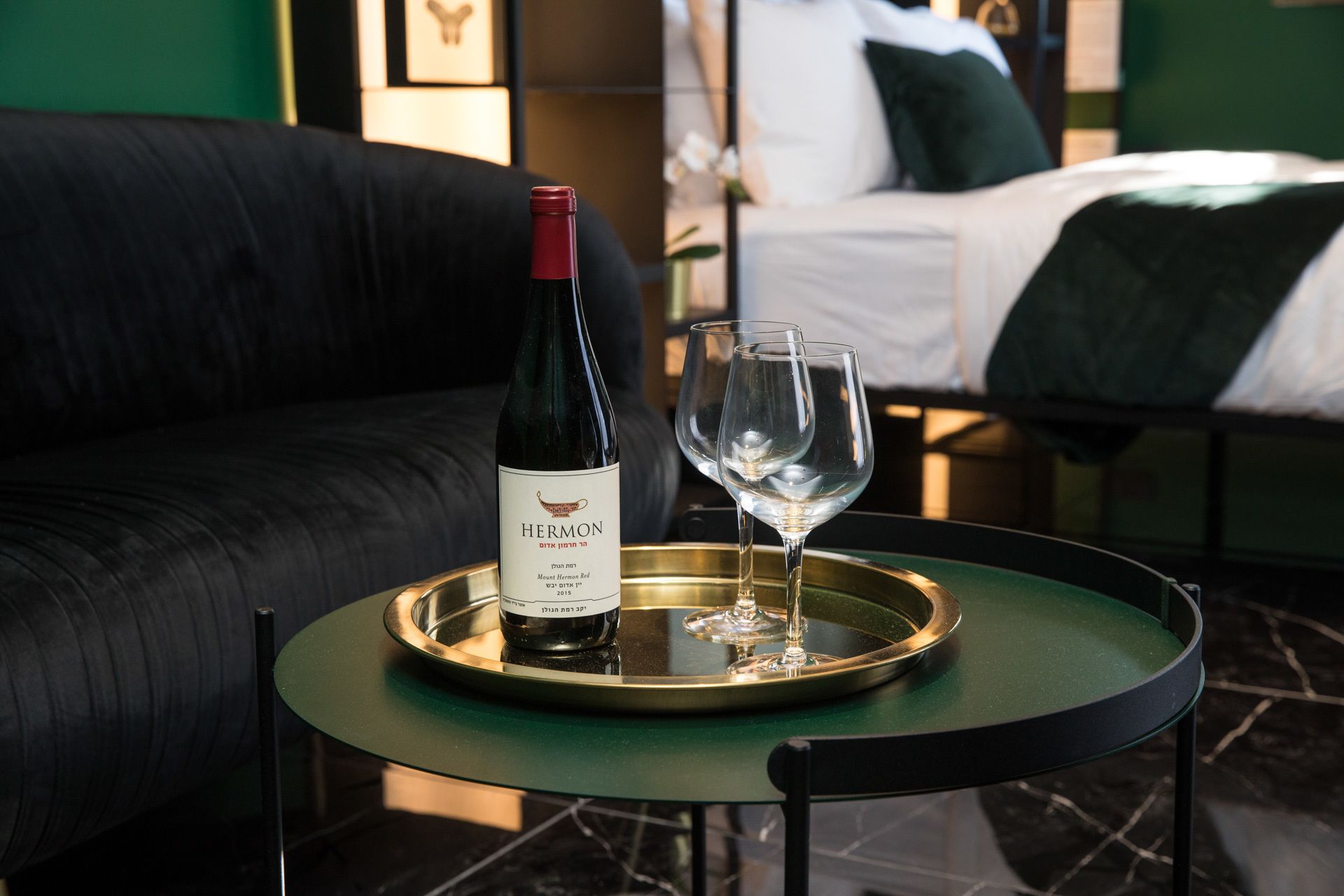 A bottle of Hermon wine on a tray with two glasses on a small round table in a room with a bed and a dark-colored sofa.
