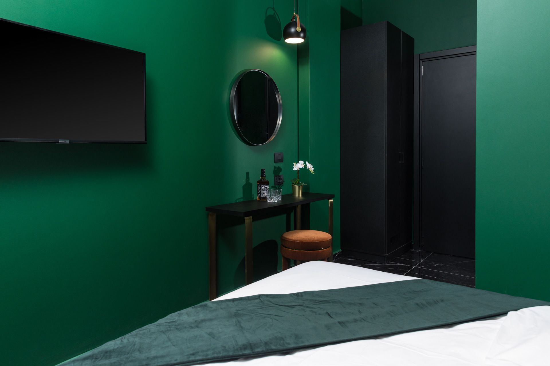 A room with green walls, a bed with a dark green bedspread, a small round mirror, a black desk with a bottle and glass, and a black wardrobe.