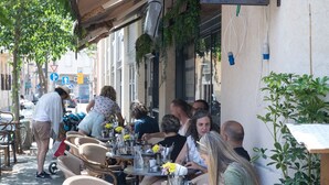An outdoor café with wicker chairs and tables, a hanging sign, and a building with a visible entrance. - Joseph Hotel TLV (Tel Aviv)