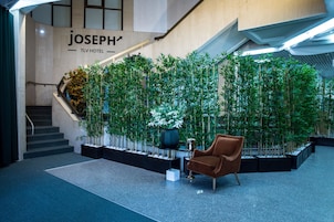 Un lobby moderno de hotel con una escalera, un letrero en la pared que dice "JOSEPH TLV HOTEL" y una zona de estar con una silla marrón.