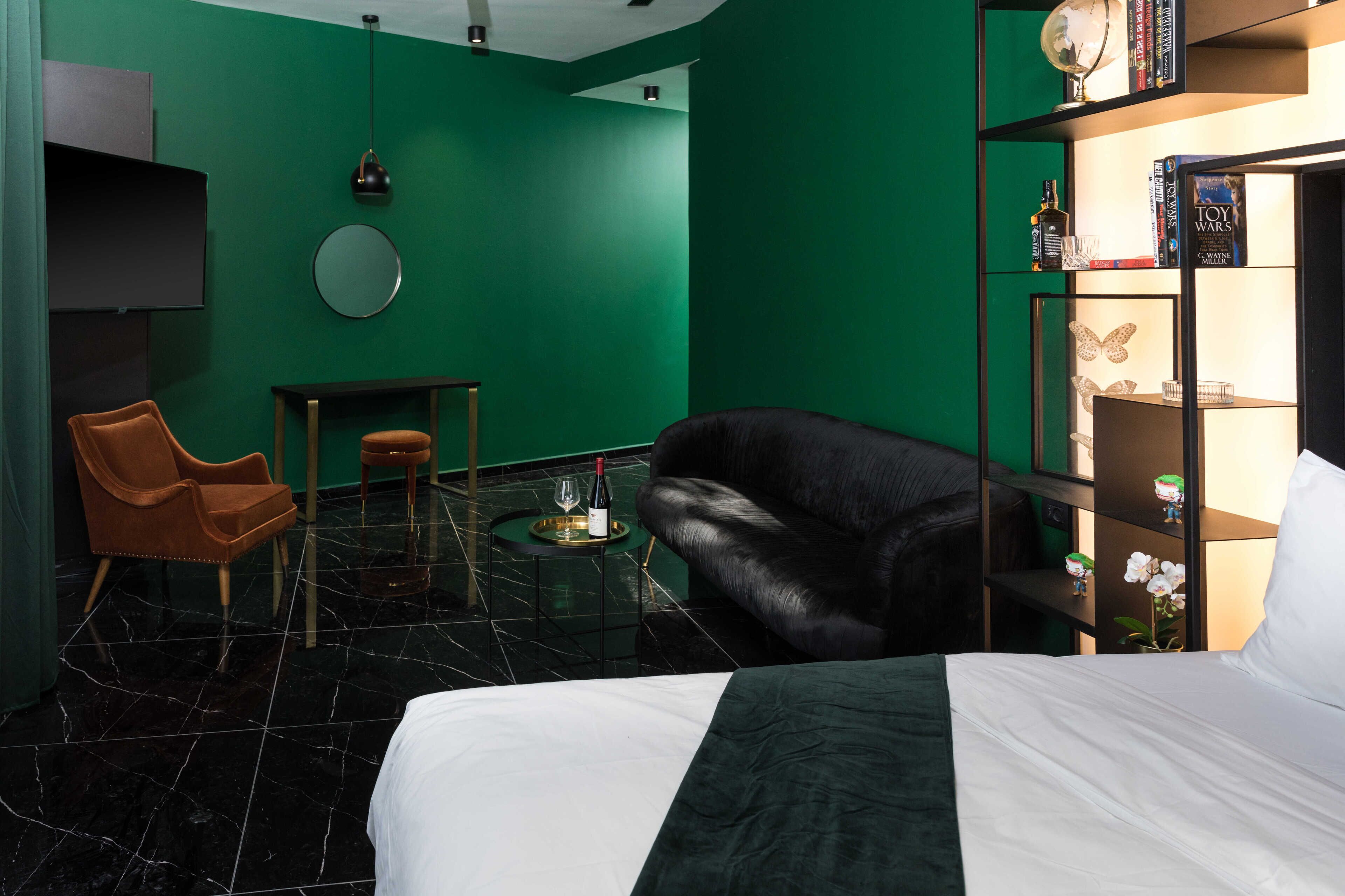 A hotel room with a bed, a leather sofa, a small table with a wine bottle, a round mirror, and a bookshelf with books and decorative items.