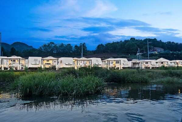 A row of RVs parked by a calm river with a backdrop of trees and hills. - Poseidon Caravan - Campsite (Gapyeong)