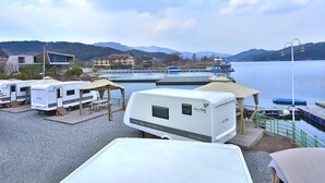 Parque de casas rodantes junto al lago, con varias casas rodantes estacionadas en un área de grava, una terraza de madera y vistas a un muelle y montañas al fondo.