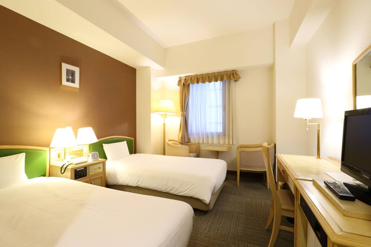 A hotel room with two beds, a desk, a chair, a TV, and a lamp.