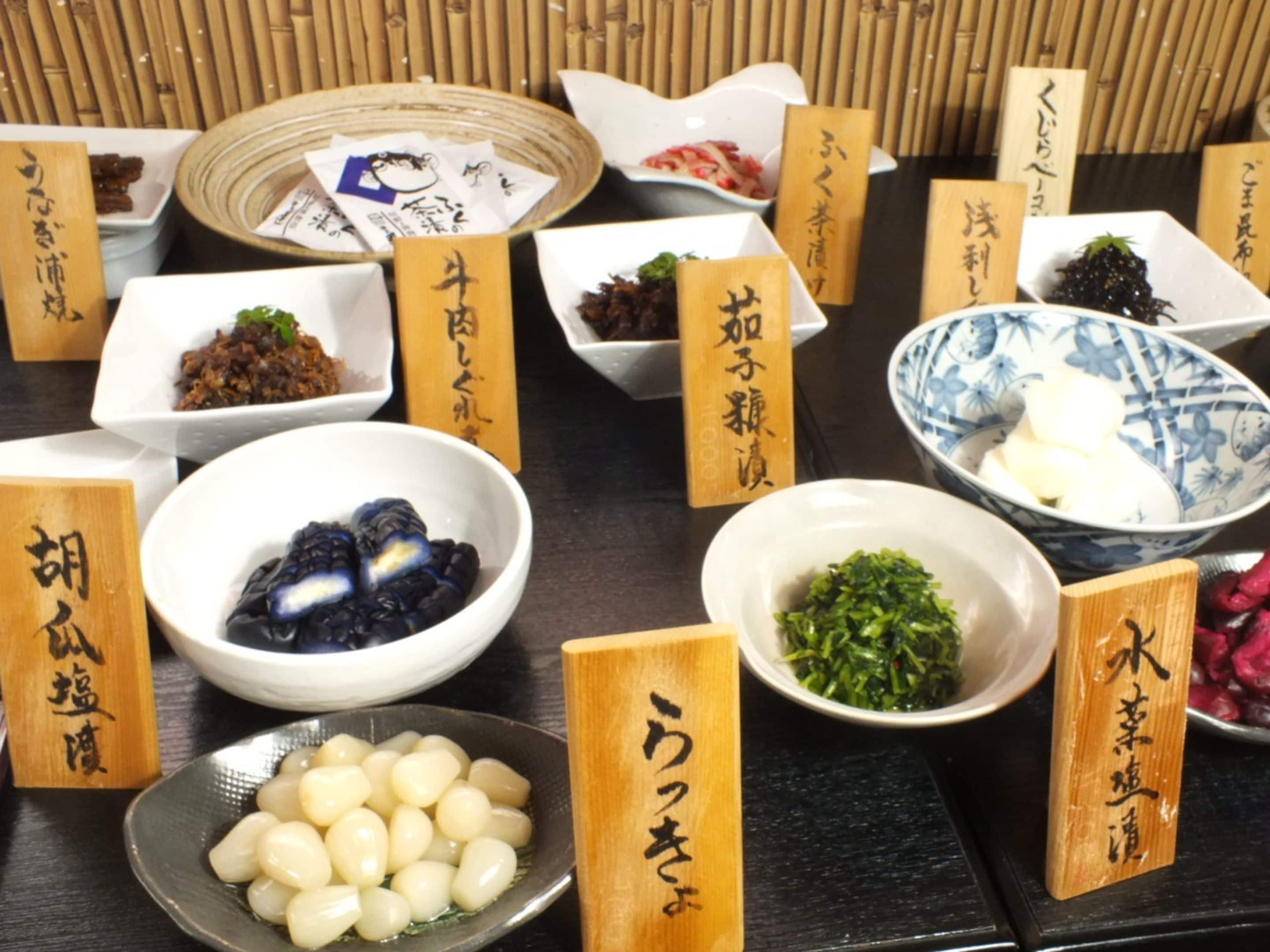 A variety of Japanese dishes with wooden labels, including pickled vegetables, dumplings, and a bowl of rice.