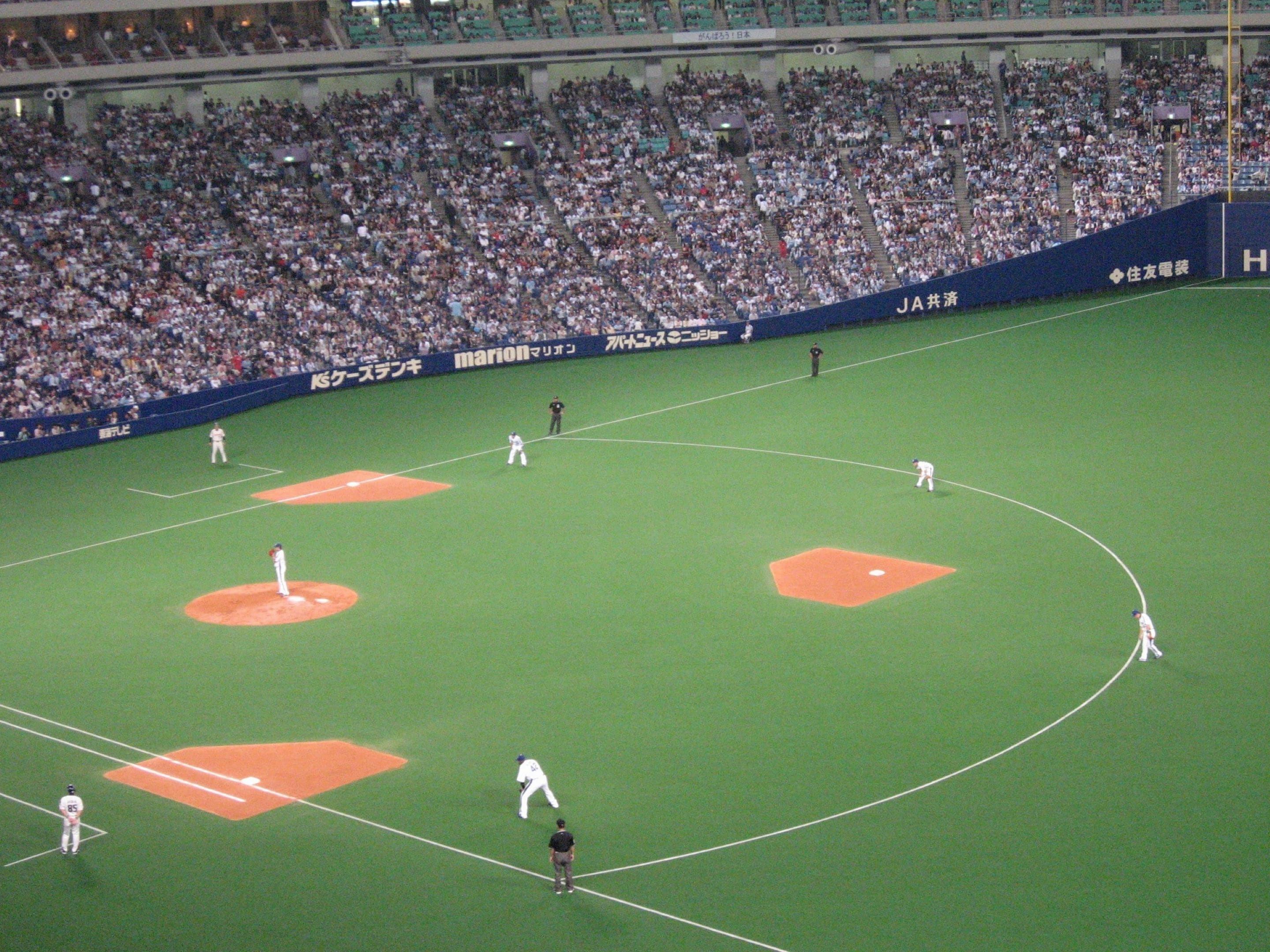 Un estadio de béisbol con varios jugadores en el campo, público en las gradas y anuncios en las paredes.
