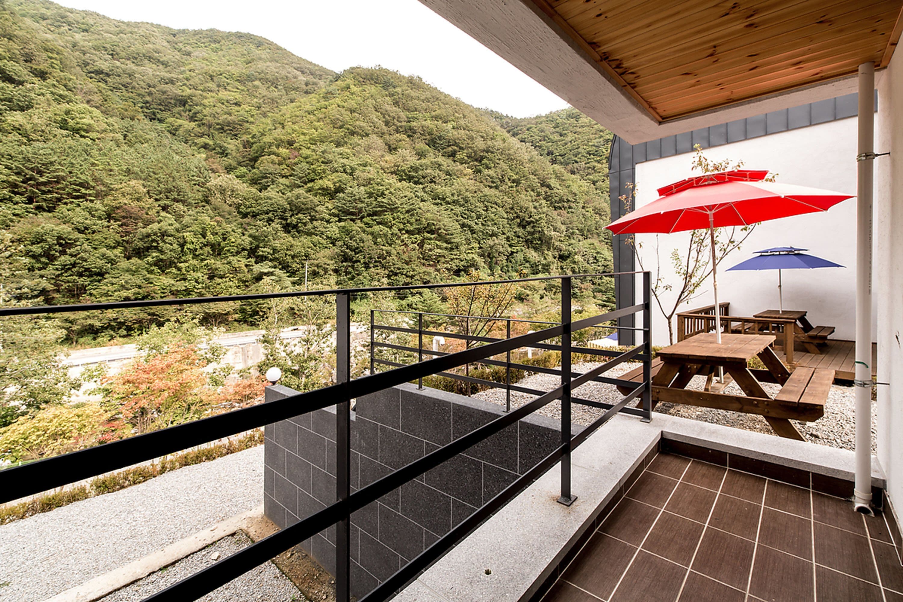 A balcony with a view of a forested hillside, a wooden table, and two umbrellas.