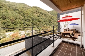 Un balcon donnant sur une colline boisée, une table en bois et deux parasols.