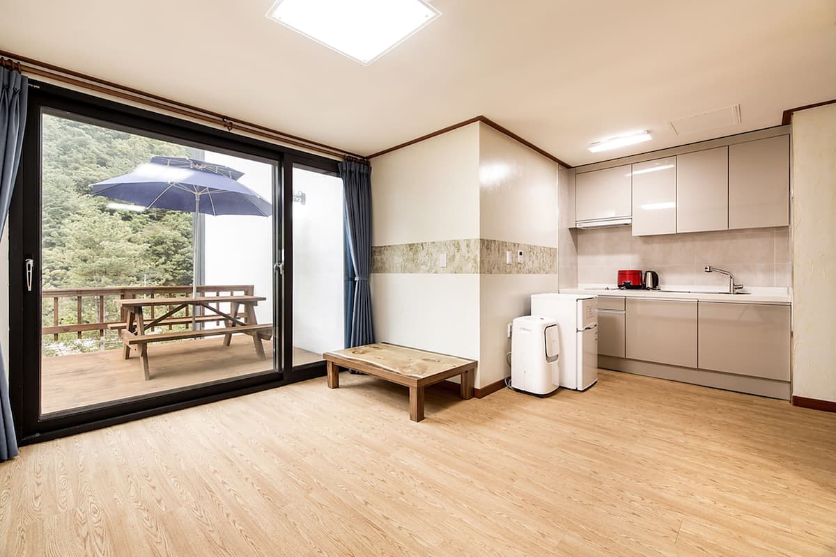 A modern kitchen with wooden flooring, a dining area with a bench and umbrella, and a view of greenery through the glass door.